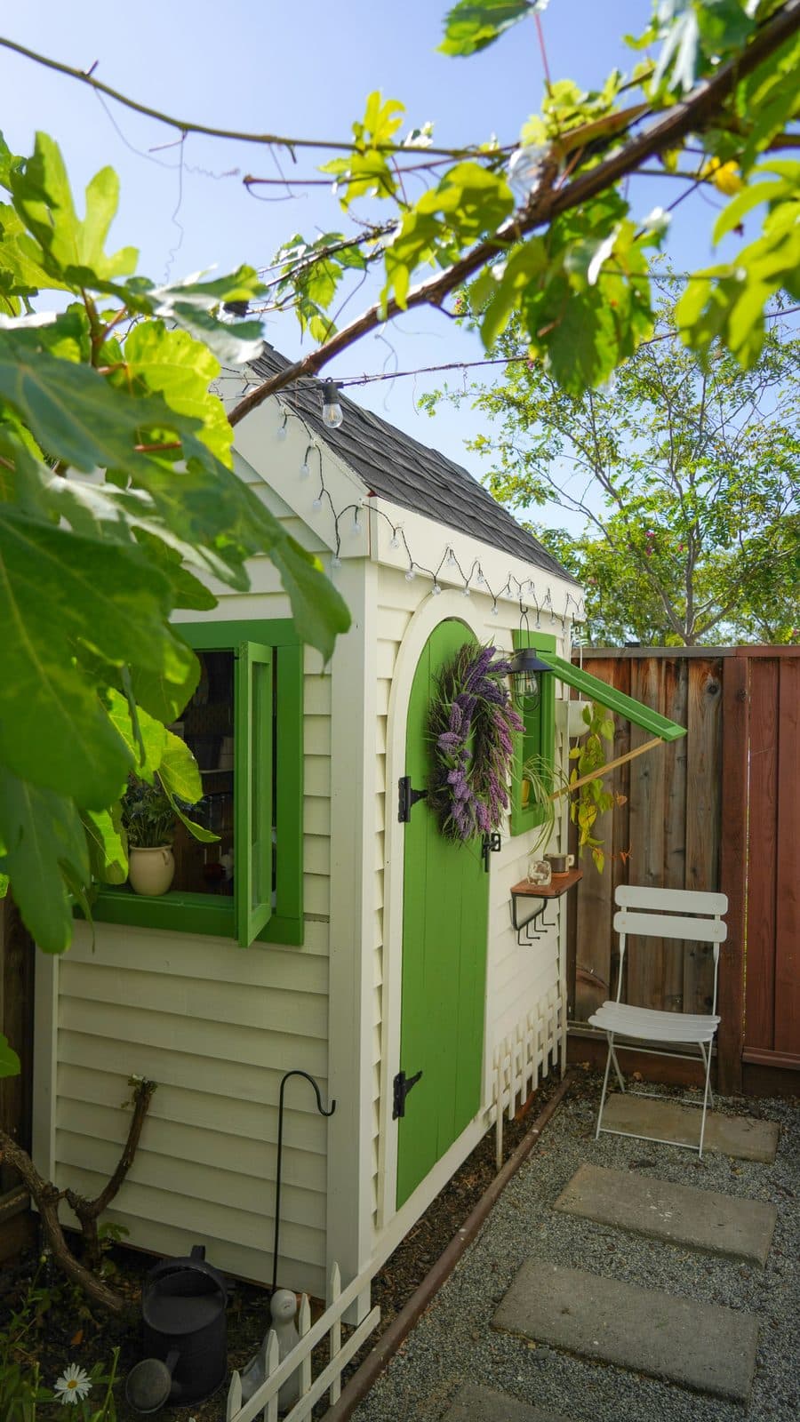 White timber garden shed with green door and shutters, set among garden foliage