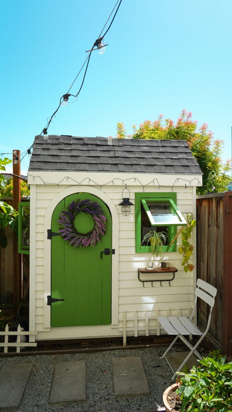 White timber garden shed with green arched door, shingled roof and string lights